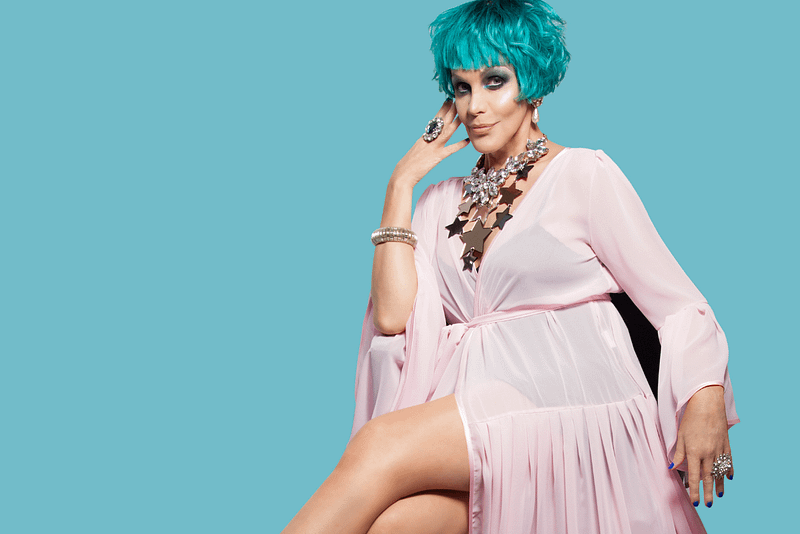 A person sits on a chair, they have blue short hair, and are wearing a light pink dress, their leg is poking out of the dress, and the background of the image is pale turquoise blue.