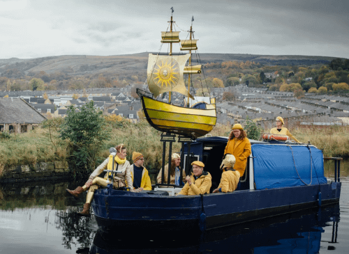 There is a boat on a canal, in the background there are hills in view, and sat on the boat are four people, and a sculpture of a yellow boat with gold sails.