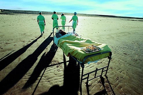Four nurses walking away from a person in a hospital bed, on a beach