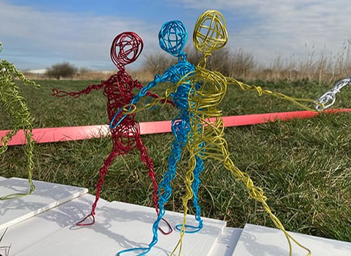 The image is set in a field, there are three wire sculptures, one is red, one blue, and one yellow. They mirror the human form and are stood in a pose that looks like they are moving.