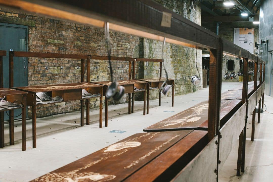 There are two rows of wooden desks sitting parallel to each other. Each one has etchings in the surface and small speakers dangling down above the desk. The background is a stone wall.