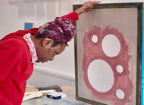 A woman is holding a piece of art work in progress. The work is made up of five circles and has a red outline. The lady has a purple headscarf on, and a red jumper.