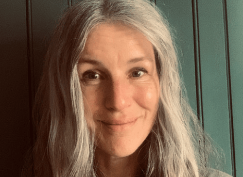 A white woman looks straight ahead. She has long grey/blonde hair and is smiling with her mouth closed.