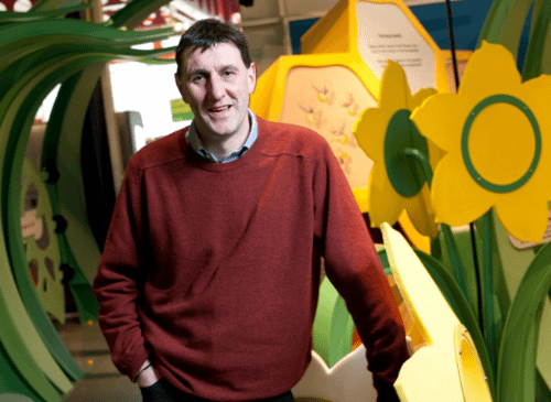 A man stands infront of a large sculptural image of yellow and green flowers. He is tall and has short hair. He is wearing a red jumper, and is smiling at the camera. He has white skin and looks like he's in his forties.