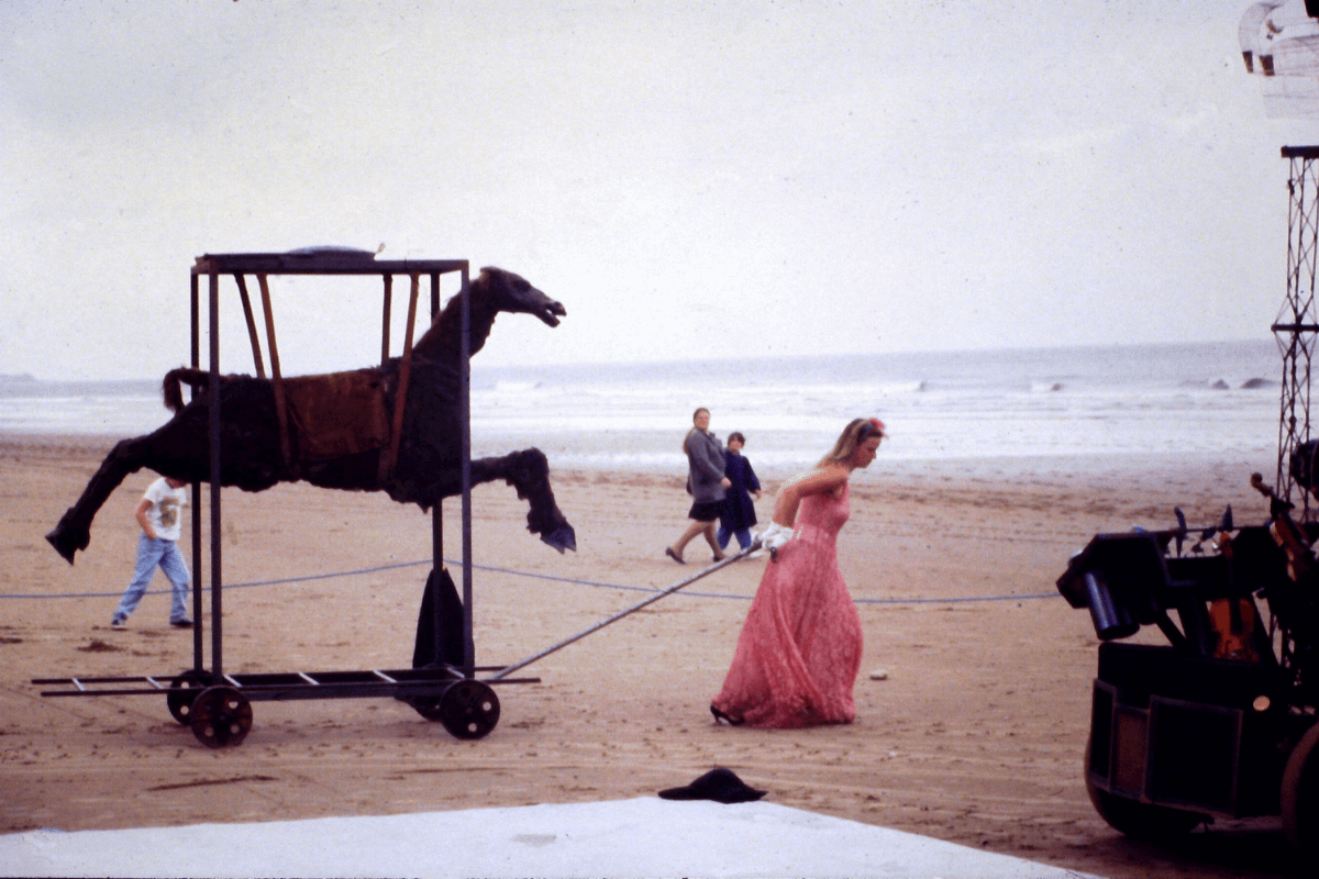 A lady in a red dress pulls a model of a brown horse along a beach.