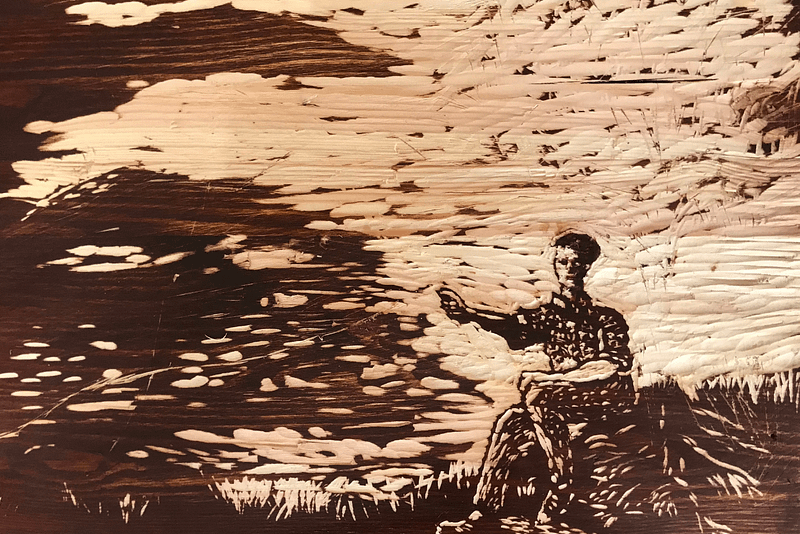 A wooden etching has contrasting foreground of dark wood, the etching creates a pale background and forms the shape of a man holding our his hand, sat next to a rive.