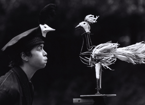 A woman has a hat on with a bird sat on top, she is blowing at the statue of a cockerel. The image is black and white.