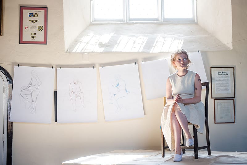 Performer Jemima Foxtrot sits under a sunlit window for life drawing class, in a white dress.