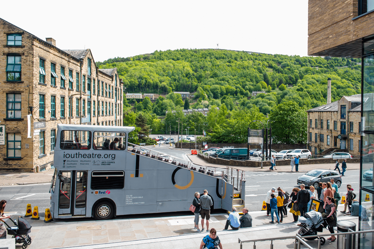 A grey bus with a diagonal section cut of out it, to make it open top is parked infront of a large mill. There are trees in the distance, and a queue of people waiting to board the bus. It's a sunny day.