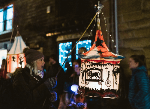 Festive lantern, held by a women wearing a wooly hat, on an outdoor parade. There are fairy lights in the background, and the atmosphere looks fun.