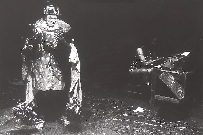 A black and white photograph, shows tow figures dressed in a large fancy dress cape and top hat. In the background there is a figure that is blurry, it appears to be sat at a desk with spots all over it's outer garment.