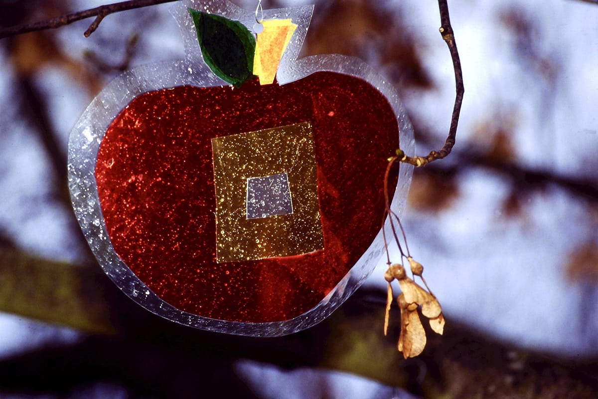 Close up photograph of a red fabric apple, with golden catkins hanging of a branch. The apple has yellow and green leaves at the top.
