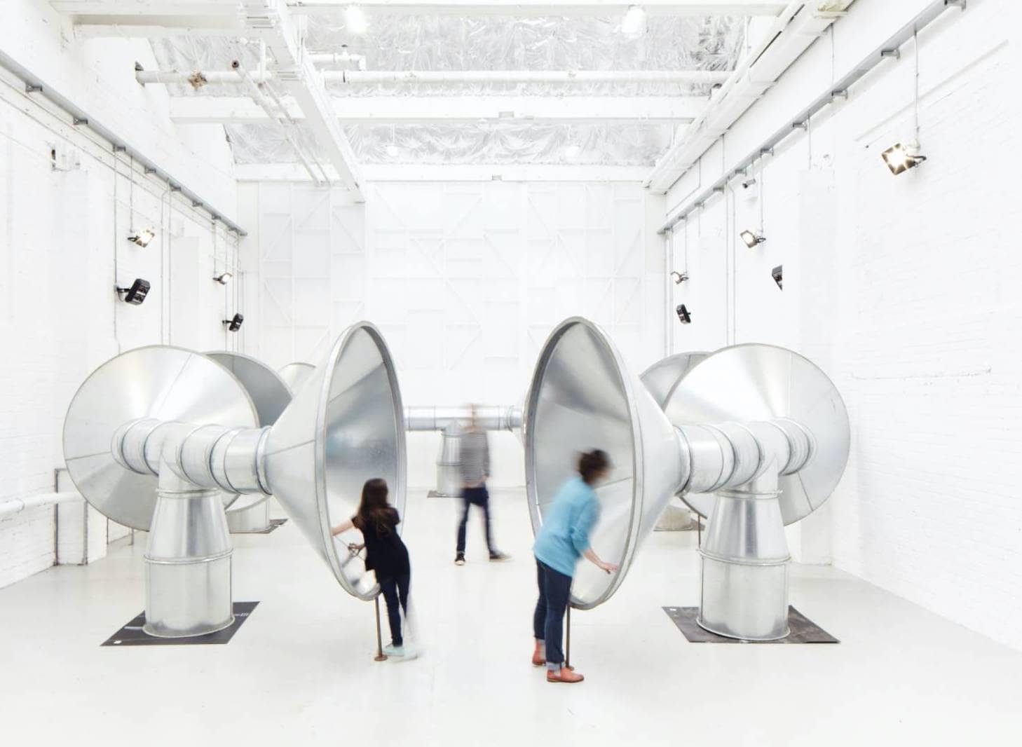 White gallery space featuring huge steel speakers with people leaning into them
