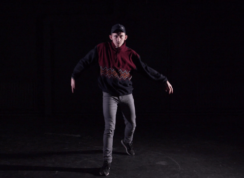 A white man stands in a dark room wearing a woolen jumper and a cap. He appears to be dancing and has his arms out in a wiggly shape.