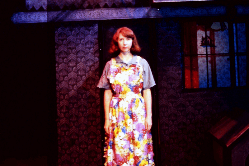 Woman is stood, looking at the camera, with red hair that sits at chin length, she has a fringe and is wearing a floral patterned dress.