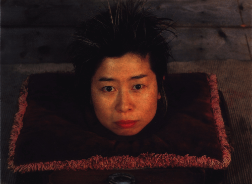 There is a velvet cushion with the head of a woman sticking out of it. The woman is looking at the camera, she has short black hair that is sticking up. She is pouting slightly, and looks confused.