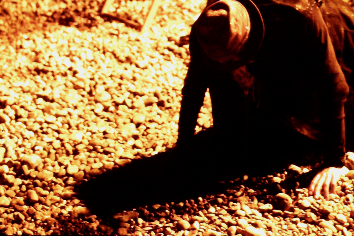 A sepia coloured image, with a man who is nealing down on a pebbly beach.