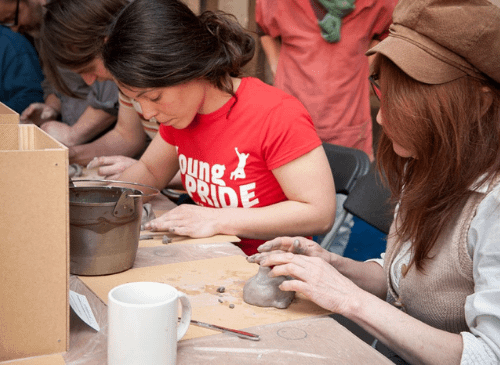 Two women sit at a desk doing craft work with clay and wood. One has hat on with ginger long hair, the other has black hair tied back and is wearing a red t shirt.