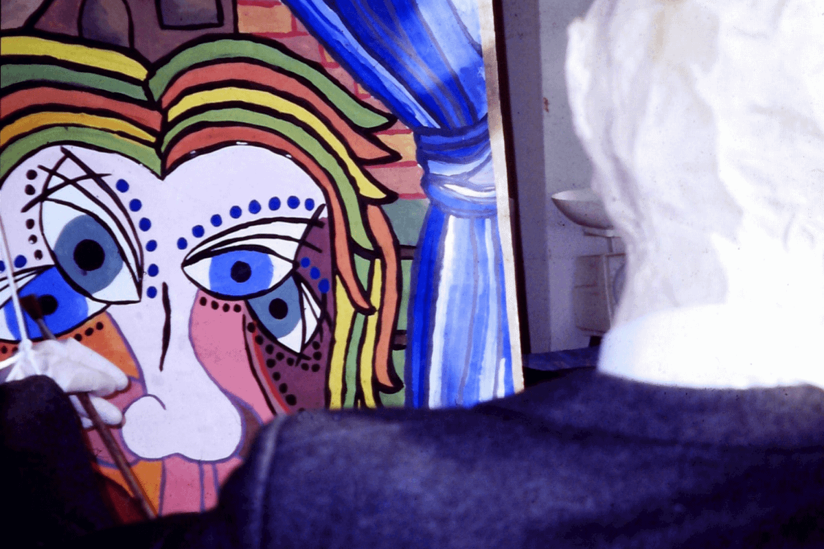 A large drawing sits in the back of a room. It's backlit and colourful and depicts a face with long black eye lashes.