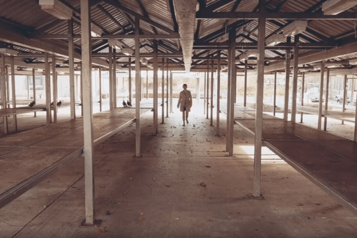 A female figure stands within a disused building. The large building is mostly metal uprights. She wears a beige trenchcoat and has white plimsoles on.