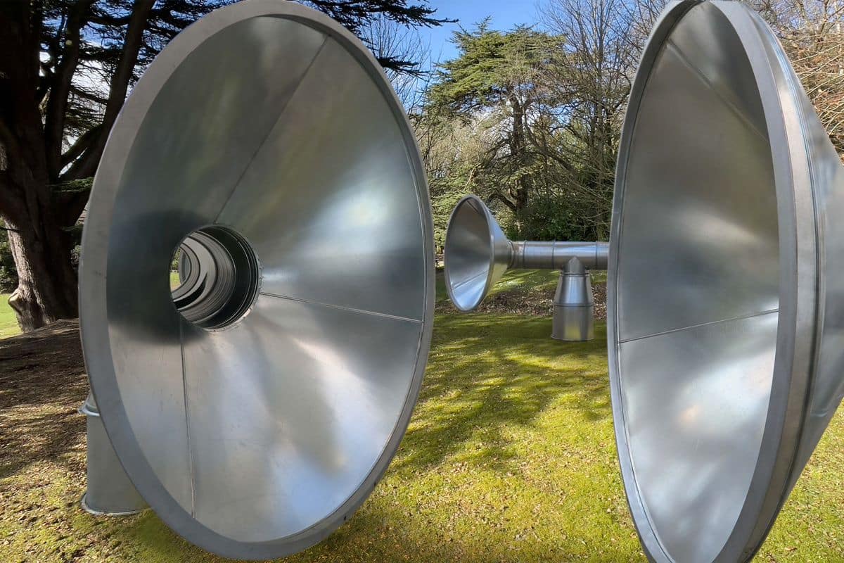 Large silver metal tubes and speakers sit in a garden with trees
