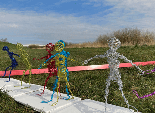 There is a row of wire figures, they stand next to each other in a field, and are white, yellow, blue, red, yellow and deep blue in colour. The sky is blue behind them.