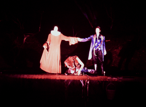 There is a women on the left, wearing a coral coloured dress, stretching out to hold the hand of a man on the right. They are both illuminated in a bright light, with a dark backdrop. There is a figure lying on the floor underneath their outstretched hands.