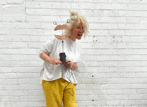 A woman stands in front of a white brick wall, holding a metal sculpture of a pair of lips. She has white skin, and blonde hair, and is wearing yellow trousers and a white t shirt.