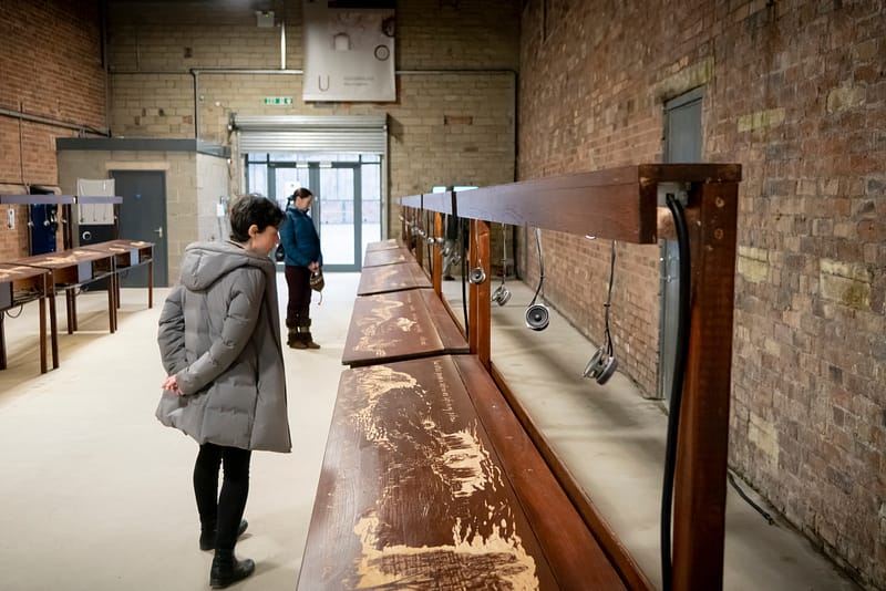 A row of wooden desks in view. There are tiny speakers hanging down which you can see from the front of the image. There are people standing looking at the desks.