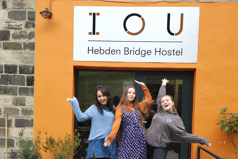 Three young women stand in front of an orange wall, with a sign above the wall that says IOU Hebden Bridge Hostel