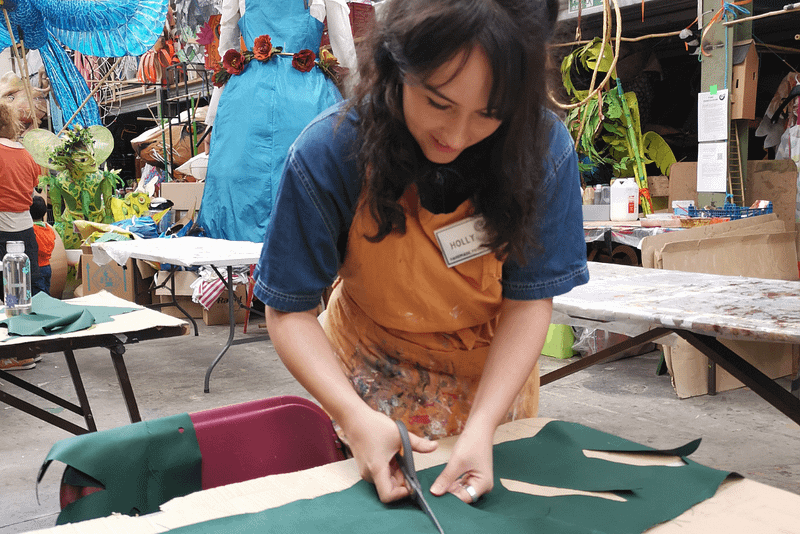 A young woman stands at a table and uses scissors to cut fabric.