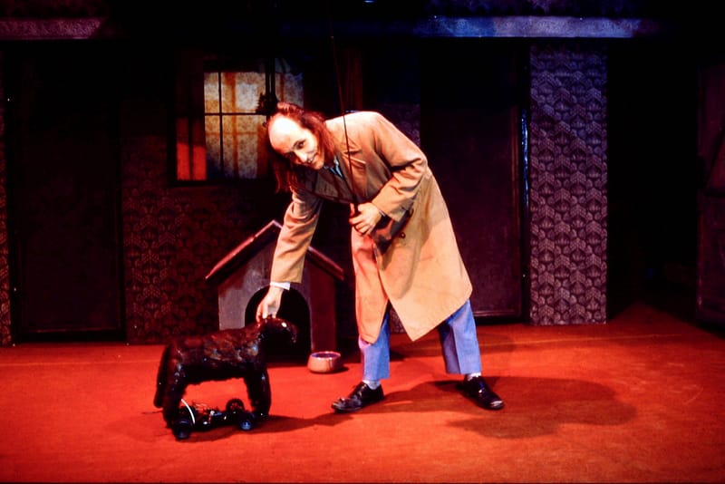 A man in a brown trench coat, reaches down to stroke a dog.