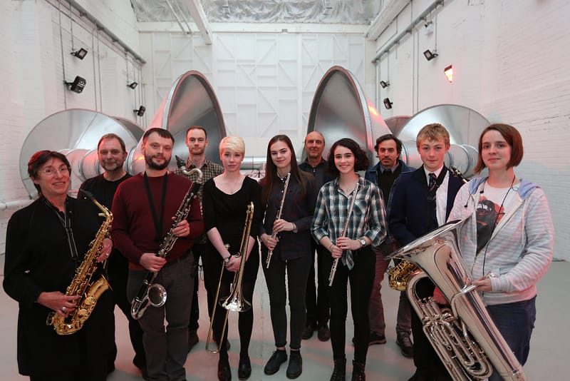 A group of 12 people stand in a row, they are holding musical instruments. In the background, there are large industrial sized tubes. They are smiling.