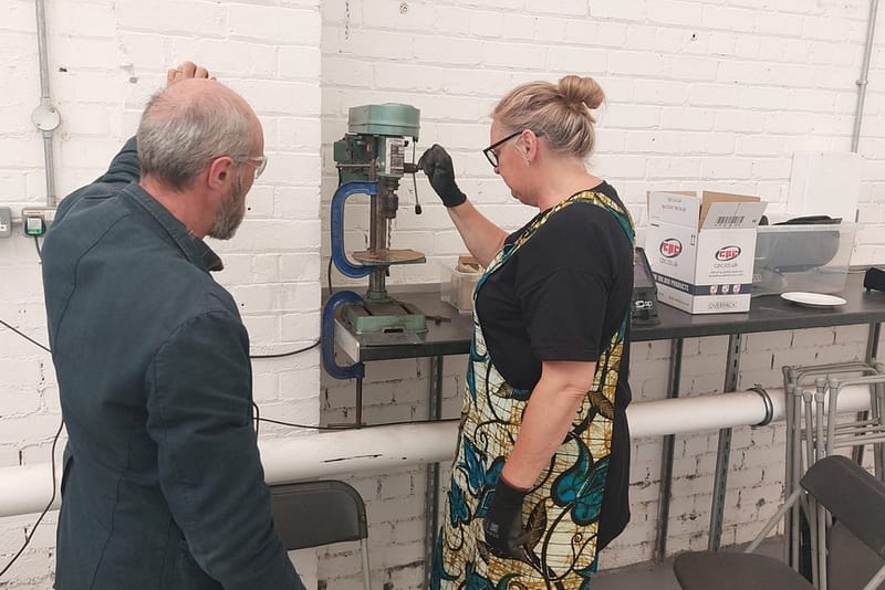 A man stands and looks at a woman as she uses a piece of machinery, the wall behind them is white. It appears that the woman is doing metal work.