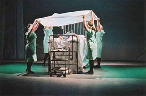 A metal hospital bed sits in the middle of an empty room. People surround the bed and hold a sheet together above the person in it.