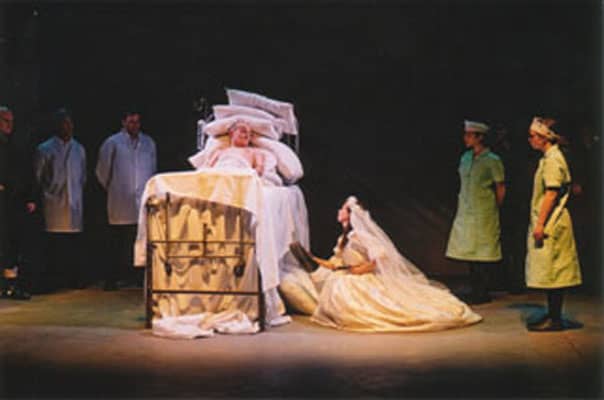 A man sits on a bed, with a lady in a bridal dress next to him.