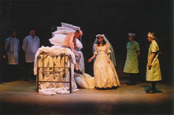 A man sits on a bed, with a lady in a bridal dress next to him.