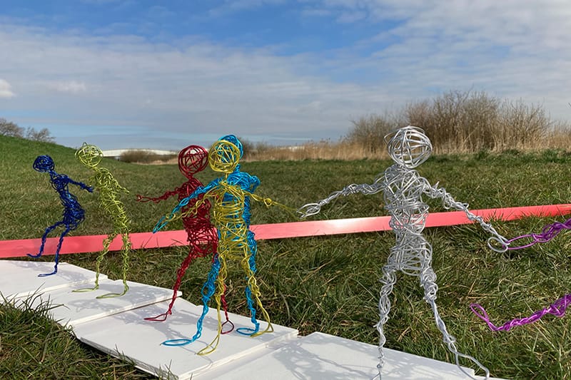 There are a row of wire figures, they stand next to each other in a field, and are white, yellow, blue, red, yellow and deep blue in colour. The sky is blue behind them.