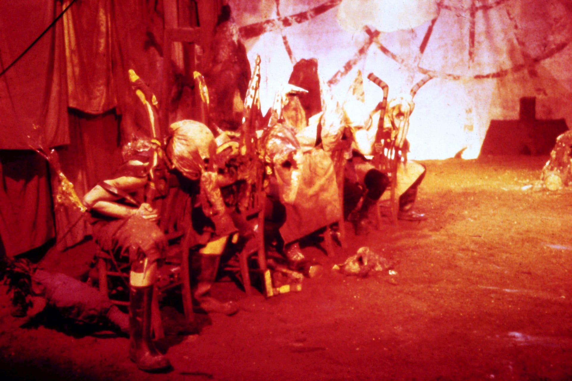 An orange light fills a room with figures sat on a chair, wearing costumes.