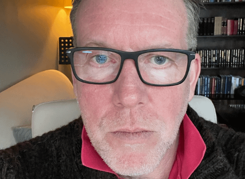 A man is looking up close to the camera. He has dark glasses on and a white beard. He has white skin and blue eyes. He wears a jumper with a red collared t shirt.