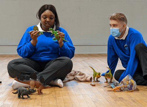 Two teenage people sit on the floor wearing blue school jumpers. There is a girl on the left who is brown skinned with dark hair holding a dinosaur. On the right is a boy who is white skinned with blonde hair, he is wearing a facemask and looking at the toy dinosaur.