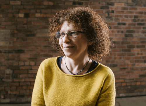 The picture shows a woman looking into the distance. She is smiling and has a yellow jumper on. She has chin length curly hair and white skin, she is wearing metal framed glasses, and has a delicate necklace on. The background is a brick wall.