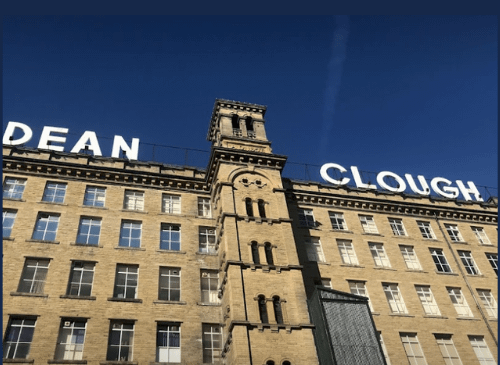 A large industrial building is viewed from the ground, the image pans up to the top of the tall building with the words 'DEAN CLOUGH' at the top.