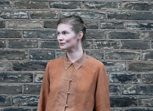A woman stands in front of a brick wall. She is wearing a rust coloured shirt and looking to the left. She has white skin and blonde hair which is tied back.