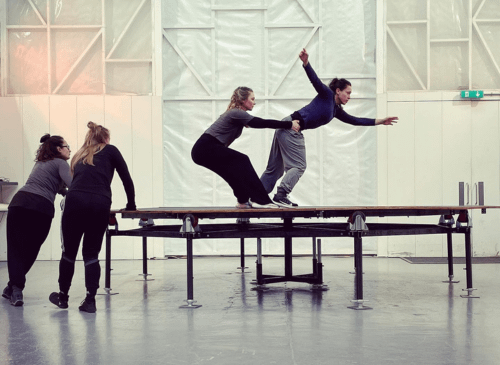 2 women dance on a large trampoline , 2 other women watch . The space is all white