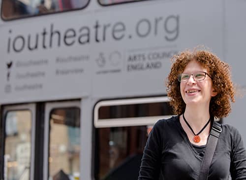 A woman with curly red hair and glasses stands in front of a white bus. The bus says IOU theatre on the front in a brown font.