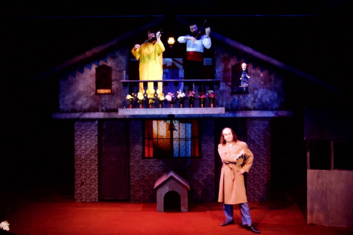 A man in a brown trench coat stands infront of a wooden house with a balcony, on the balcony are two men who are playing an instrument. One is dressed in yellow, the other has a white shirt with dark trousers.
