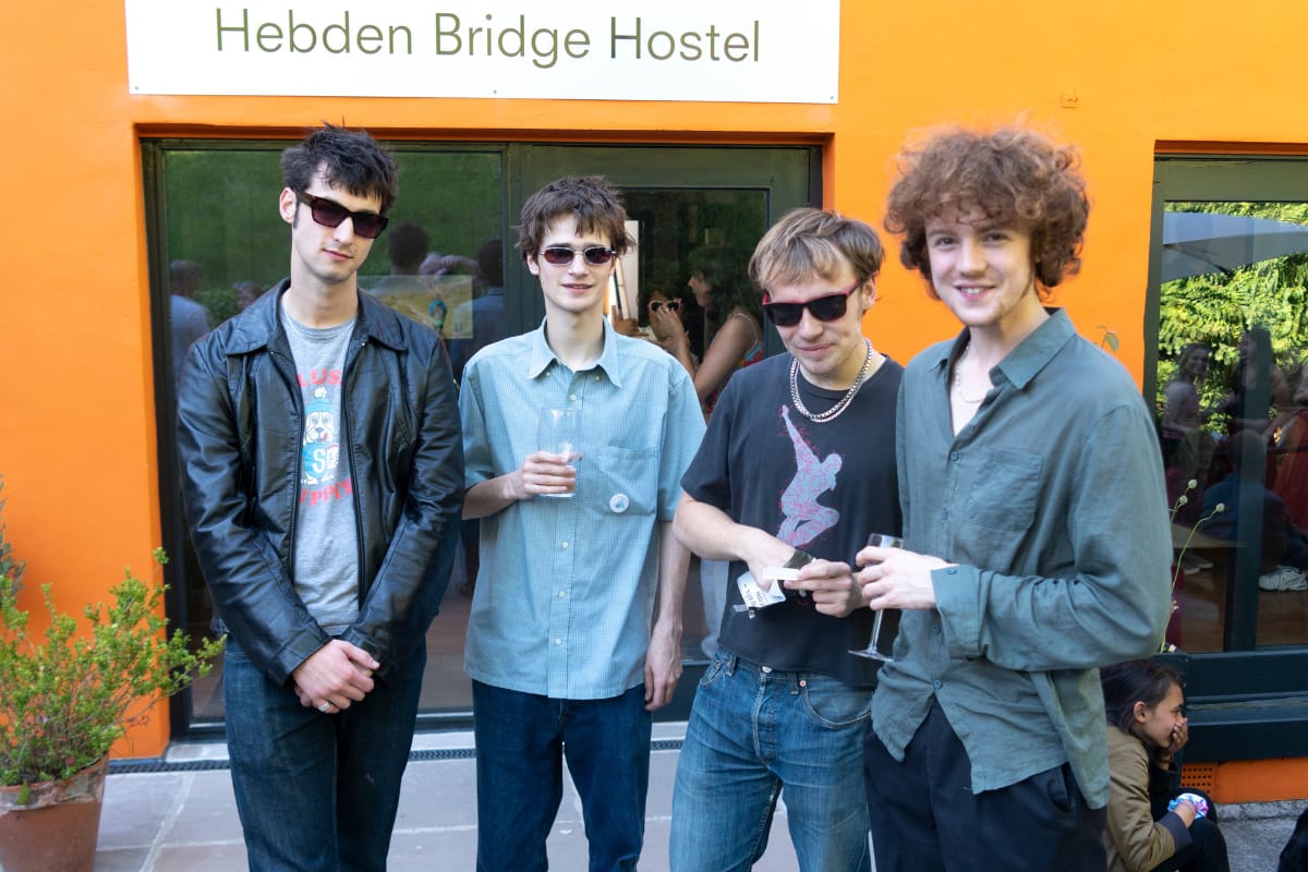 Four young men stand in front of an orange wall. There is a sign above them which has the words 'Hebden Bridge Hostel' written on a sign. They all have sunglasses on, apart from the one on the far right who isn't wearing sunglasses, he is holding a glass of white wine in his hand.